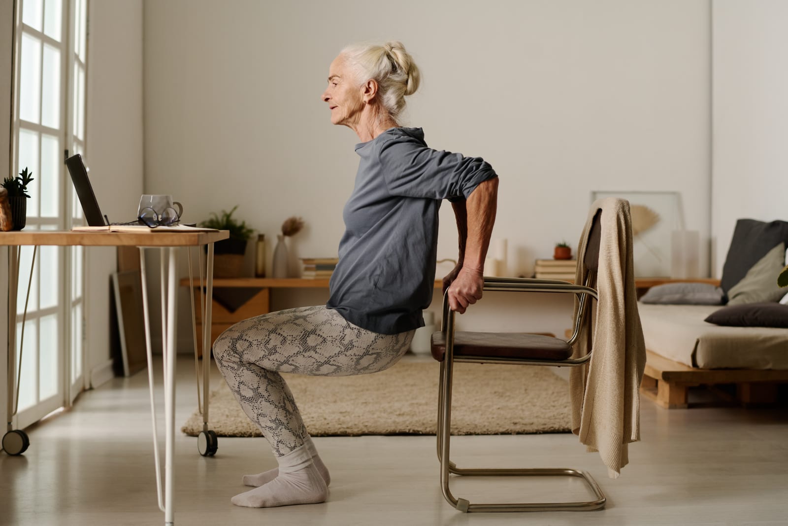 Older woman rising from a chair at home, hand resting on the chair for support